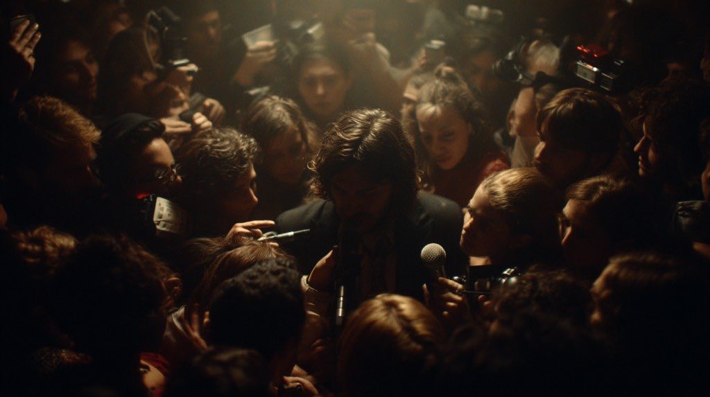 Man surrounded by a dense press crowd with cameras and microphones under overhead light